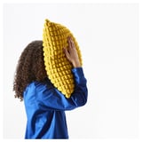 Person holding large, bright yellow, textured cushion. Bright blue clothing, simple design.