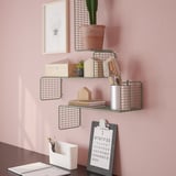 Wall-mounted grid shelves with various items: a potted plant, brushes, books, and a clipboard with a calendar. Personalized organization on pink wall.