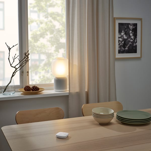 Dining room with SYMFONISK remote, lamp, pinecones, and stacked dishes. Minimalist chairs and butterfly art.