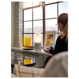 Woman works by bright window in simple room with desk and lamp.