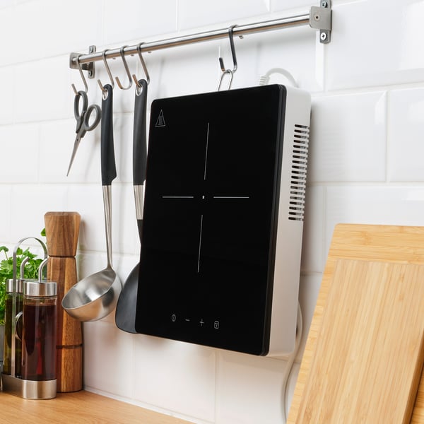 Modern kitchen with a black portable induction hob on a white countertop. A wooden cutting board and cooking utensils hang above it.