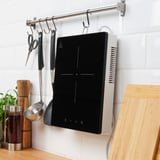 Modern kitchen with a black portable induction hob on a white countertop. A wooden cutting board and cooking utensils hang above it.