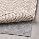 A folded beige cotton rug, perfect for living rooms or under tables, shown with an anti-slip rug underlay.
