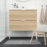 Modern bathroom with wooden vanity, glass shower, grey rug, beige stool & towels