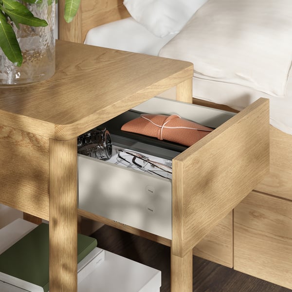A wooden bedside table with an open drawer showing neatly folded clothes and a magazine. Part of the TONSTAD series, offering storage solutions.