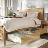 Bedroom with light wood bed, open drawer, pillows, plant, coffee, nightstand lamp, bright windows.