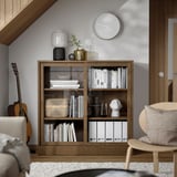 Wooden TONSTAD bookcase with glass doors, books, decor, clock, guitar.