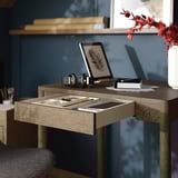 Desk with open drawer, stationery, tablet, headphones, red flowers. Shelves with books and a framed picture.