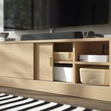 Wooden tv stand with sliding doors, showing a white device on shelves, a wicker basket, and a soundbar on top near a black tv screen.