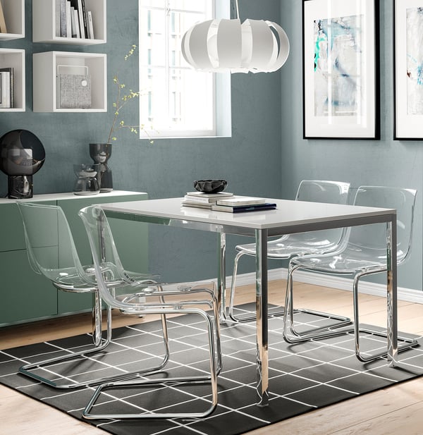 Modern dining set with acrylic chairs, white table, and pendant light on a gray rug. Decorated shelves and window in background.