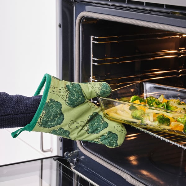 Person placing tray of vegetables into oven, wearing green oven mitts with a broccoli pattern.