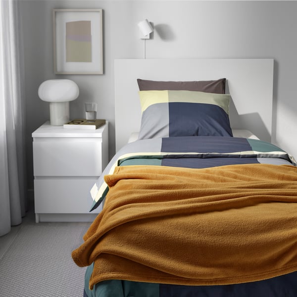 Mustard bedspread, multicolored pillows, white headboard, lamp, books. Minimalist, cozy room.