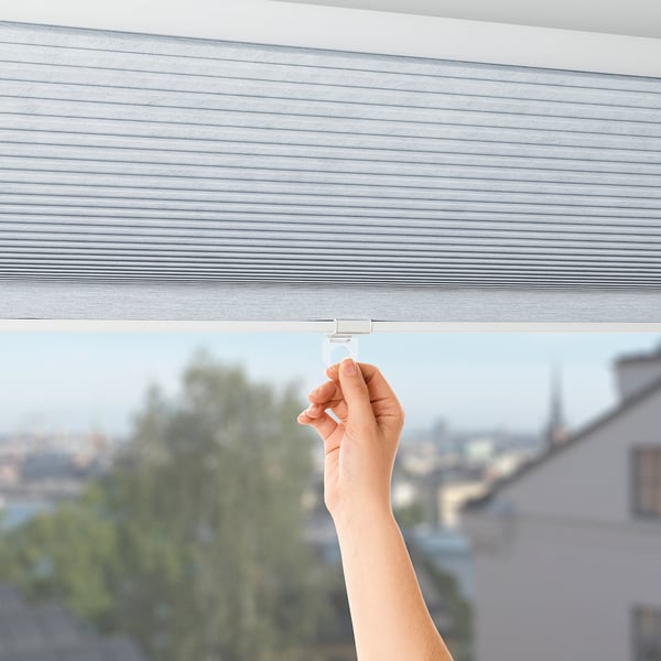 Hand raising white blind cord, revealing city view, demonstrating light control and cord safety.