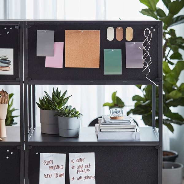 Black office storage unit with open shelves displaying plants, books, and a potted succulent. Includes a corkboard with various notes and pins.
