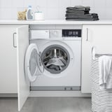 White 2-in-1 washer/dryer behind open cabinet, surrounded by towels & supplies, with striped basket nearby.