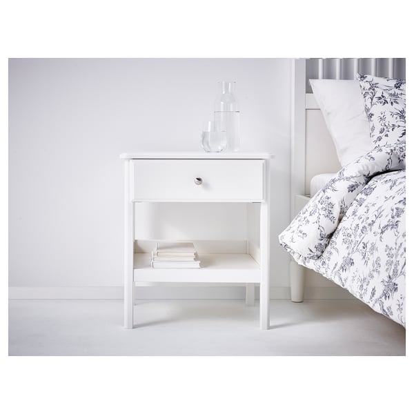 White bedside table with drawer, books, vases, next to bed with floral bedding.