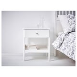 White bedside table with drawer, books, vases, next to bed with floral bedding.