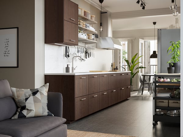 Modern kitchen with dark wood cabinets, a white marble backsplash, stainless hood, and large windows. A cozy sofa and indoor plants enhance the space.