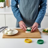 Person slices boiled eggs using green egg slicer on wooden board. Other tools and sliced eggs nearby.