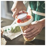 Two hands using an orange and white UPPFYLLD jar opener to twist off a lid from a glass jar filled with chickpeas.