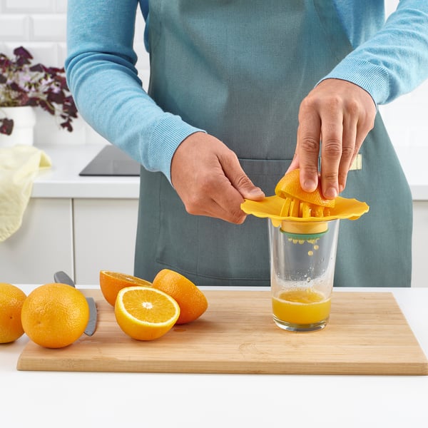 Person squeezing orange juice using a yellow lemon squeezer onto a glass on a wooden chopping board. Whole and halved oranges nearby.