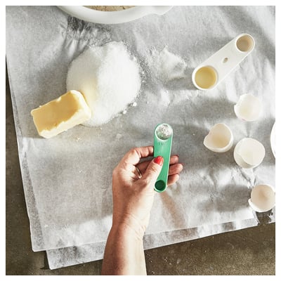 A hand holding a green measuring spoon near baking ingredients.