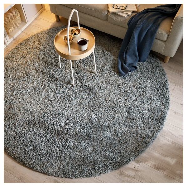 A round, grey UTREDNING area rug on a wooden floor, with a side table and couch nearby. The rug has a soft, shaggy texture.