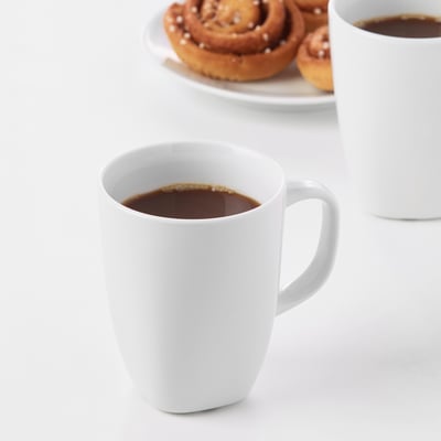 Two white ceramic mugs filled with coffee on a white table, accompanied by a plate of cinnamon buns.