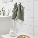 Green VALLASÅN towels hang on white-tiled bathroom wall, with a counter holding cotton swabs, toothbrushes, and a plant.