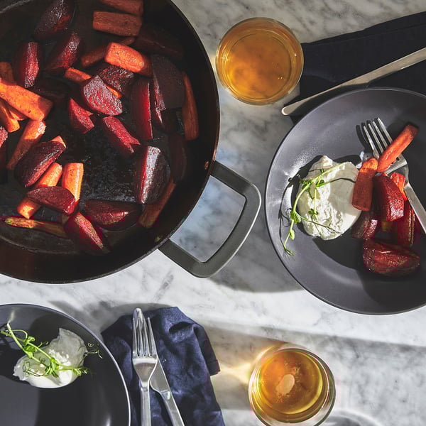 A meal is spread with roasted vegetables, pan with VARDAGEN carbon steel, on grey plates alongside forks and drinks.