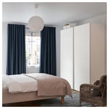 Minimalist bedroom with light pink bedding, white bed frame, dark blue curtains, hanging white light, wicker chair, black rug, and white closet.