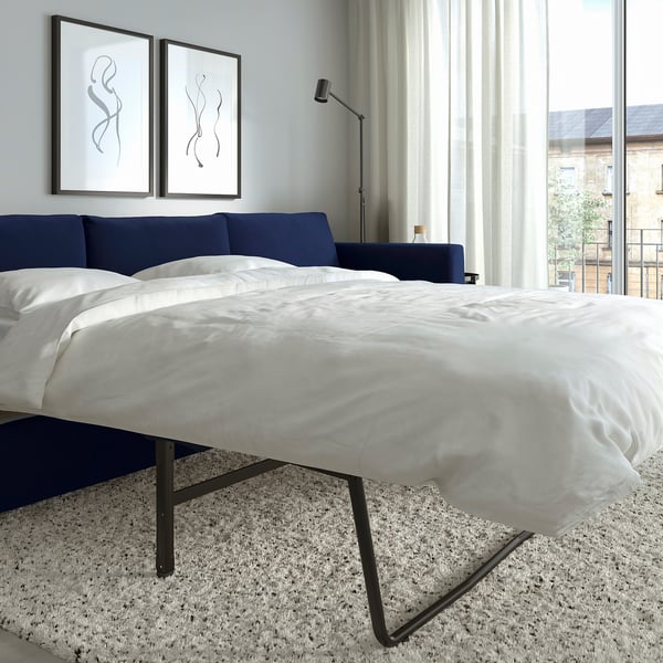 Modern bedroom with large bed, white linens, navy blue headboard, abstract wall art, floor lamp, and city view from windows.