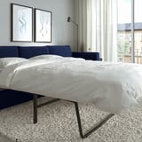 Modern bedroom with large bed, white linens, navy blue headboard, abstract wall art, floor lamp, and city view from windows.
