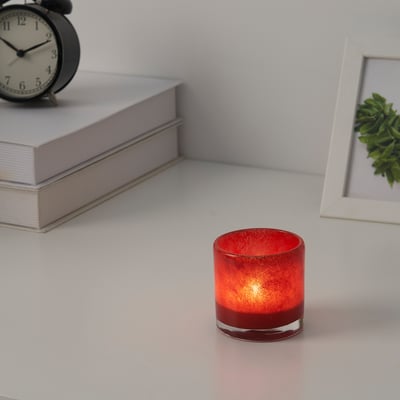 Red candle holder on table, lit, with books, clock, plant picture. Cozy.