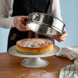 A person removes a springform pan, revealing a baked cake dusted with powdered sugar on a cake stand.
