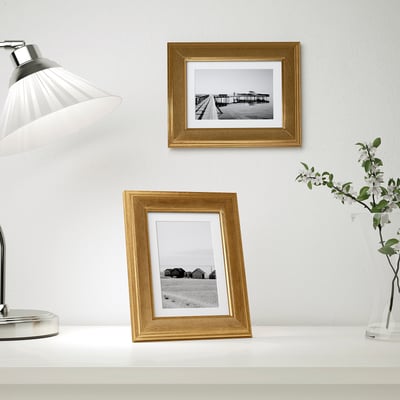Two gold frames: one on wall, one on table, with bw photos, lamp, vase.