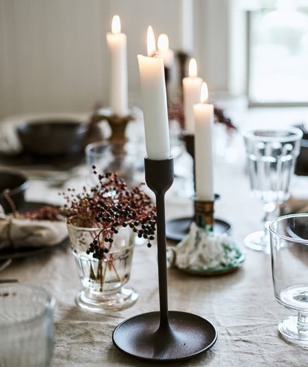 เทียนที่จุดแล้วอยู่ในเชิงเทียนสีดำและทองเหลืองบนโต๊ะปูผ้าลินิน แก้วน้ำ และกิ่งเบอร์รี่ในแจกัน