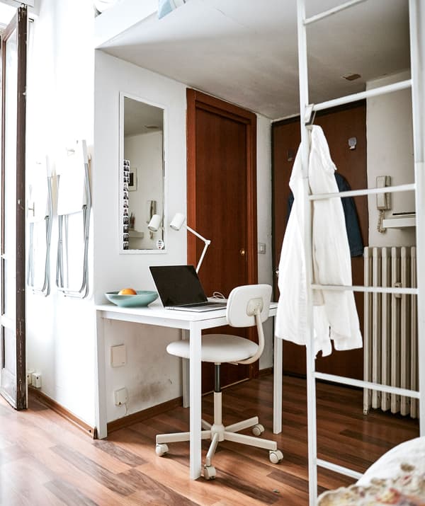 โน้ตบุ๊ควางอยู่บนโต๊ะที่อยู่ในพื้นที่ใต้เตียงชั้นลอย มีเสื้อเชิ้ตสีขาวแขวนอยู่ที่บันไดสีขาว