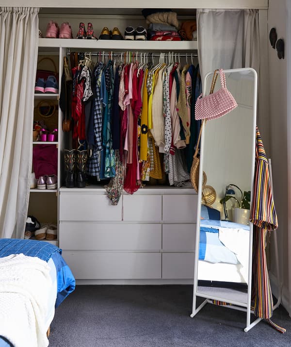 เสื้อผ้าแขวนอยู่บนราวและรองเท้าวางอยู่บนชั้นเหนือตู้ลิ้นชักสองลิ้นชัก มีม่านอยู่ทั้งสองข้าง และกระจกแบบตั้งลอย