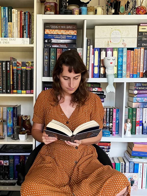 אישה בשמלה ארוכה יושבת בכיסא וקוראת ספר. על הקיר מאחוריה יש מדף ספרים עם שלל ספרים.