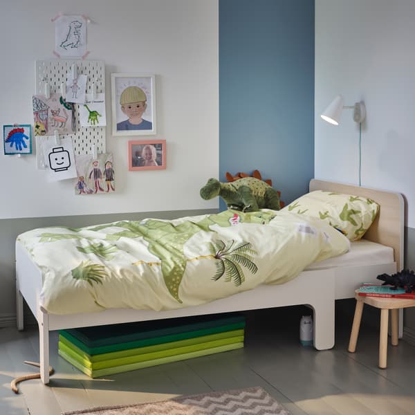 공룡 패턴 침구와 부드러운 공룡 장난감이 놓여 있는 화이트/자작나무 길이조절 침대. 그 아래에는 그린 색상의 놀이매트가 접힌 채로 놓여 있습니다.