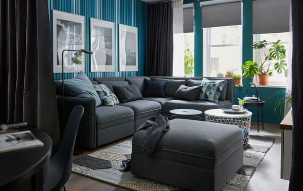 ห้องนั่งเล่นที่อากาศถ่ายเทพร้อมหน้าต่างขนาดใหญ่ โซฟาเข้ามุม ผนังและผ้าม่านสีเข้ม รวมทั้งโต๊ะกลางขนาดเล็กและต้นไม้จำนวนหนึ่ง