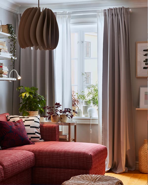 โคมไฟแขวนเพดานสีเบจ โซฟาสีแดง สตูลวางเท้าทำจากหวาย ผ้าม่านสีขาวและสีเทา และต้นไม้สีเขียวบนขอบหน้าต่าง