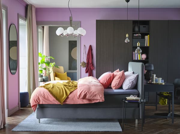 라일락 톤 벽, 라이트브라운레드 색상의 침구, 옐로 스로우, 블랙브라운 옷장, 그레이 색상의 디반침대로 컬러풀하게 꾸민 침실.