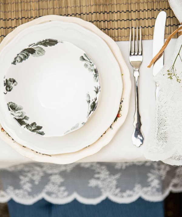 จานลายดอกไม้วางซ้อนกันบนจานรูปทรงต่างกันสองใบ มีเครื่องใช้บนโต๊ะอาหารบนผ้าปูโต๊ะสีขาว