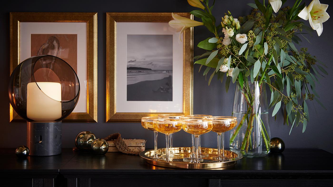 유리 꽃병에 담긴 꽃장식과 벽에 걸린 두 개의 금색 사진 액자 앞에 샴페인잔이 놓인 트레이가 있어요.