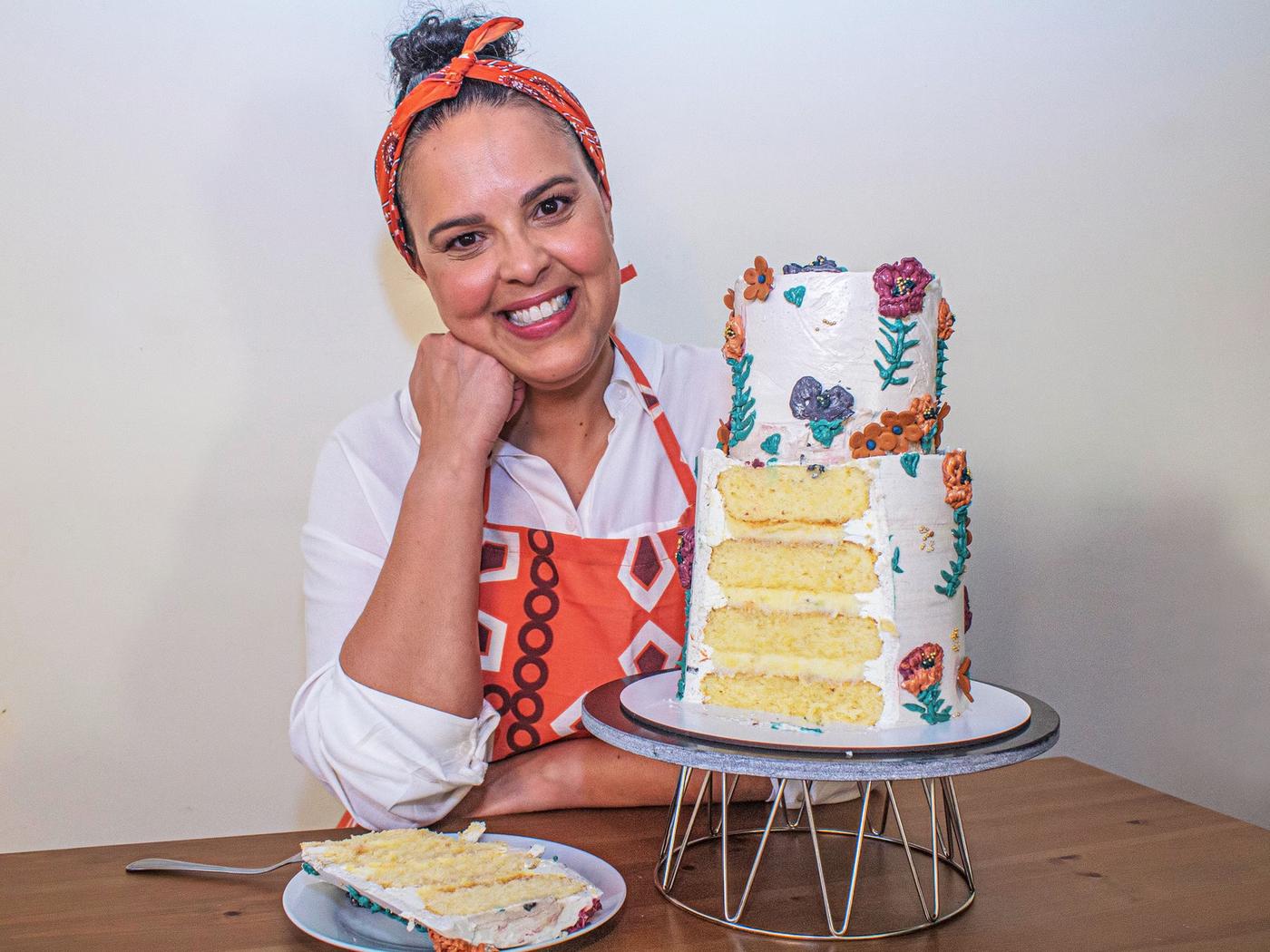 אישה מחייכת בסינר יושבת ליד שולחן, לצד עוגת שכבות גבוהה עם קישוטים יפים.