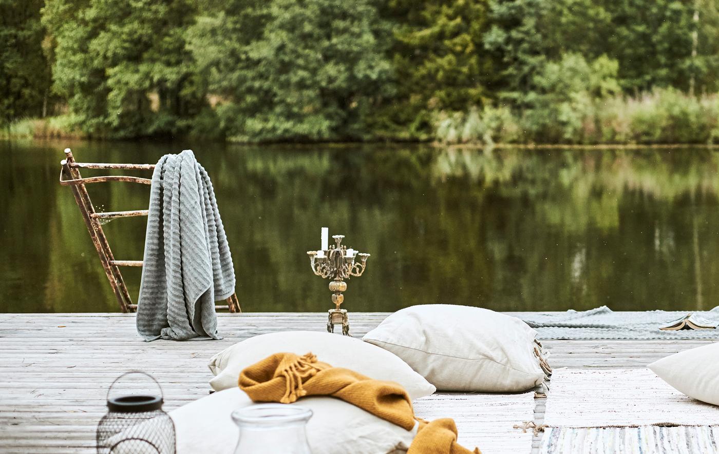 ท่าเรือริมทะเลสาบ มีหมอนอิงและพรมปูบนพื้นสไตล์ชนเผ่าเบดูอิน ผ้าเช็ดตัวสีเทาแขวนอยู่บนบันไดที่ขึ้นสนิมถัดจากเชิงเทียน