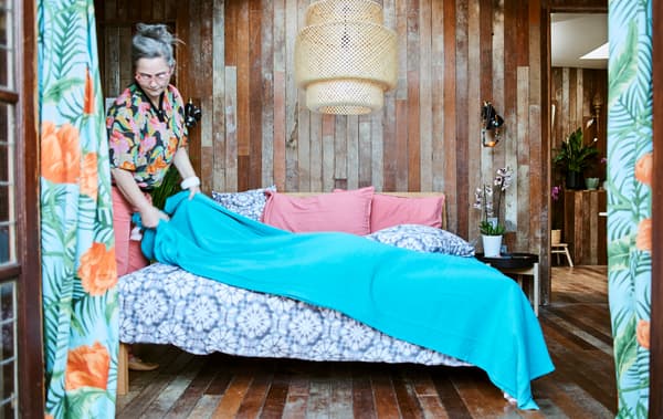 ผู้หญิงคลุมเตียงด้วยผ้าคลุมสีฟ้า เครื่องนอนมัดย้อมสีม่วงในห้องนอนผนังไม้ที่มีโคมไฟสานแขวนเพดานขนาดใหญ่