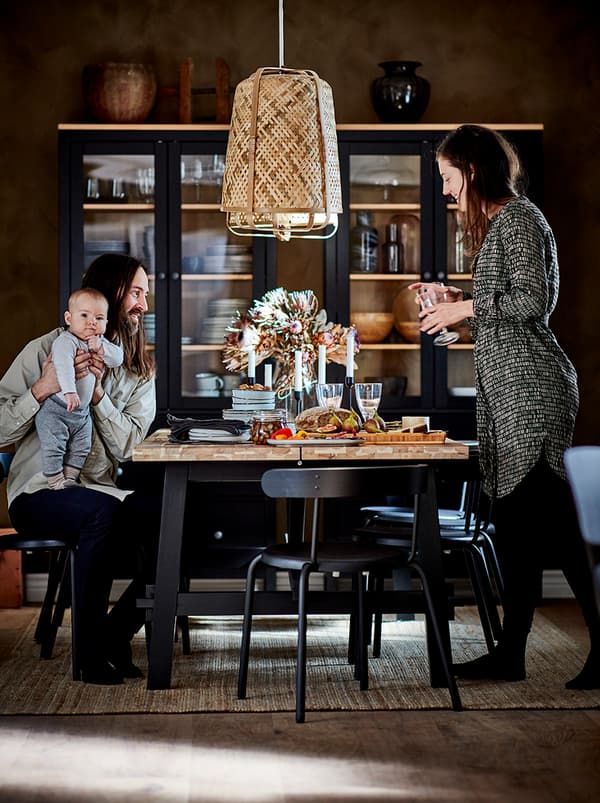 สามีภรรยาพร้อมลูกน้อยอยู่ด้วยกันที่โต๊ะอาหาร ตู้บานกระจกสีเข้มพร้อมเครื่องใช้สำหรับโต๊ะอาหารตั้งอยู่ติดผนังที่ห่างออกไป